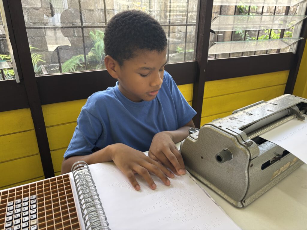 Op de foto is een jongen met kroes haar te zien. Hij heeft zijn ogen dicht en zit achter een tafel. Zijn handen rusten op een pagina met braille. Naast hem op de tafel staat een Perkins brailleschrijfmachine.