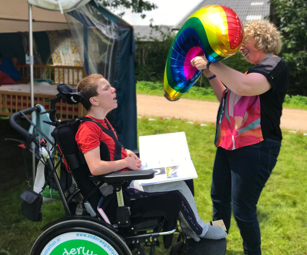 Links op de afbeelding zit een jongen in een rolstoel. Achter hem is een stukje van een voortent te zien. Rechts op de pagina staat een vrouw. Zij houdt regenboog-ballon ter hoogte van haar gezicht.